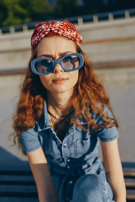 Lifestyle woman wearing modern boho-western denim street style with oversized sunglasses and a red patterned headband in warm film color tones outdoors.
