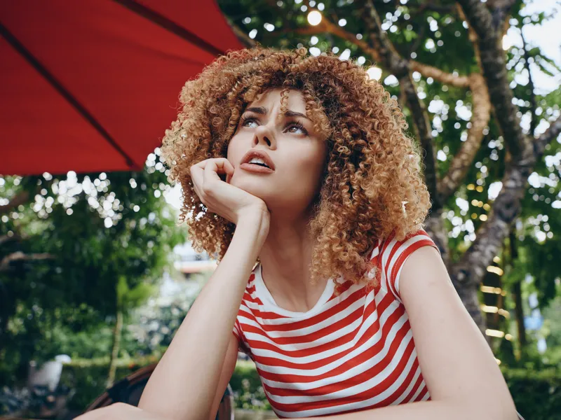 Young woman with curly hair rests her chin on her hand, wearing a striped red and white shirt, under a bold parasol in a lush garden, daylight portrait.