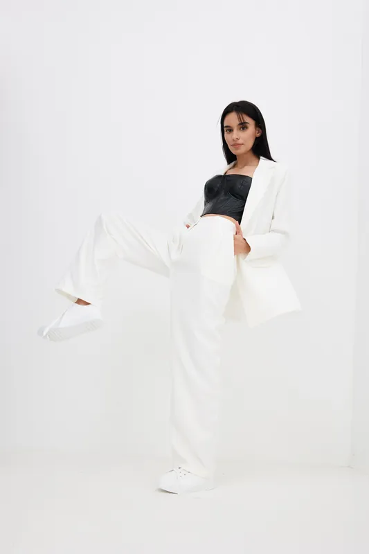 Young woman posing confidently in stylish white suit and black top with casual white sneakers against minimalist white background, showcasing modern fashion and elegance.
