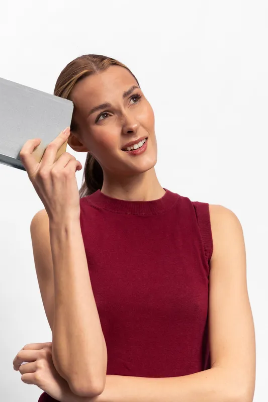 Young woman in red sleeveless top holds gray book near head, looking thoughtful. Studio shot with white background. Concept for thinking, education, and inspiration.