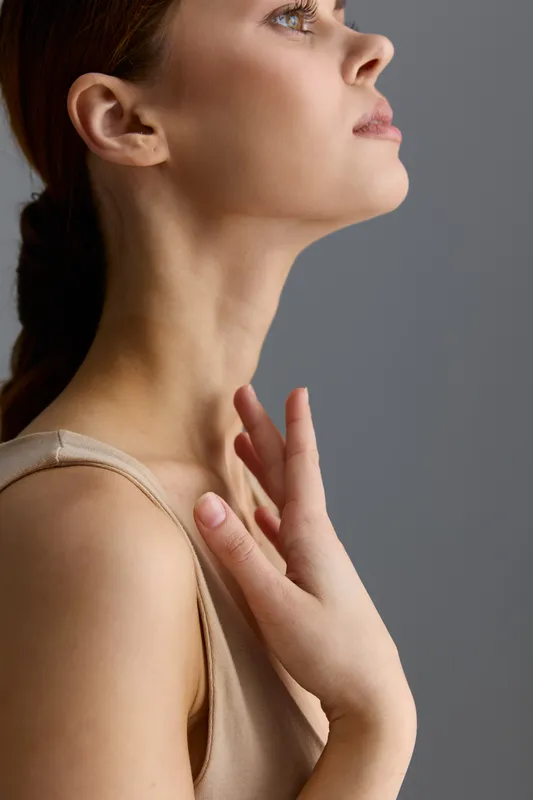 Profile of a woman with clear skin touching her neck gently, showcasing natural beauty and calm expression against neutral gray background with soft lighting.