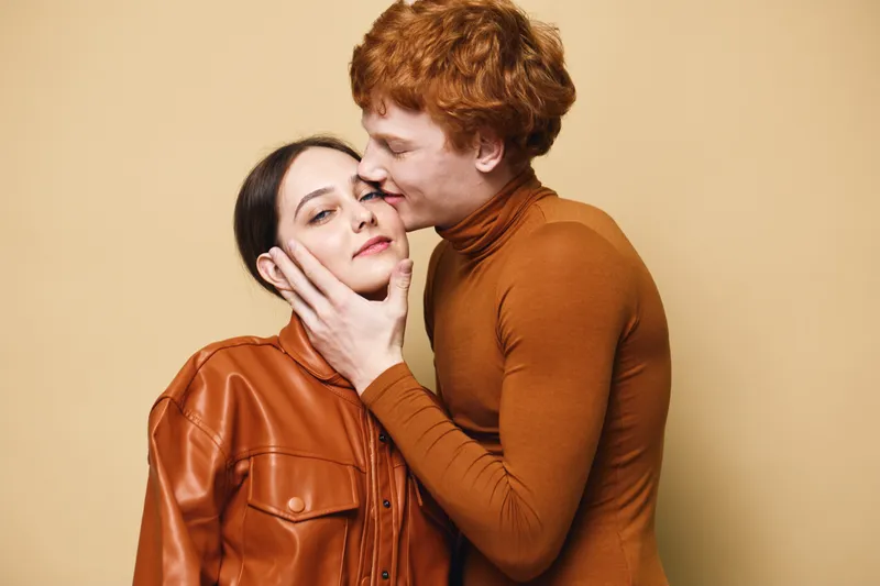 Couple with affectionate gesture, man gently kissing woman on cheek with hands on face, showing intimacy and love in warm tone clothing against beige background.
