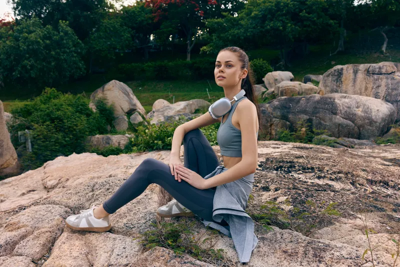 Young woman practicing yoga outdoors on rocky terrain, wearing sportswear and headphones, surrounded by greenery and autumn colors, conveying calmness and fitness