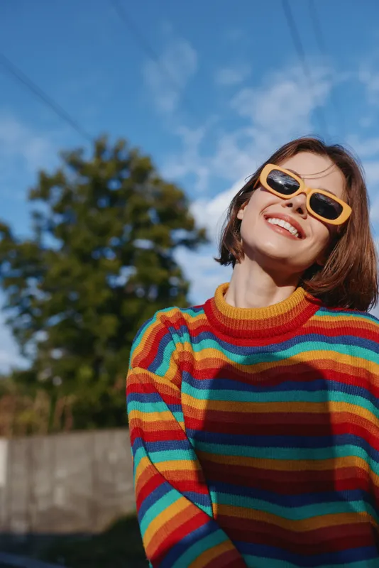 Woman smiling in yellow sunglasses and colorful striped sweater, outdoor portrait with happy casual vibe, playful fashion, short hair, bright sunny mood, urban background and relaxed pose.
