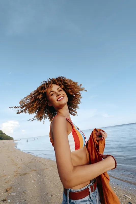 Smiling young woman standing on sandy beach beside ocean, holding a brown dog on a sunny summer day, curly hair in wind, blue sky background, outdoors relaxation near shoreline with calm waves