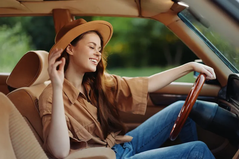 Joyful young woman in a brown outfit, enjoying a sunny ride in her vintage car, exuding confidence and happiness with nature in the background.
