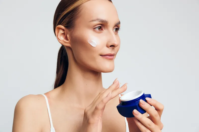 Young woman applies face cream on cheek holds blue jar with moisturizer looks thoughtful isolated on white background skin care beauty routine concept natural makeup.
