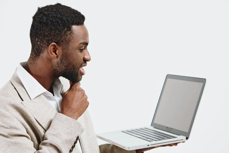 Thoughtful man looking at laptop screen, working or studying at home, casual beige sweater, focused expression, modern workspace, clean background. People lifestyle concept