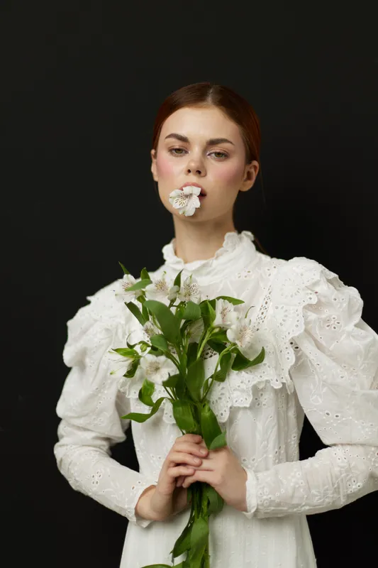 Elegant young woman in a white vintage dress holding flowers, conveying tranquility and beauty against a dark background