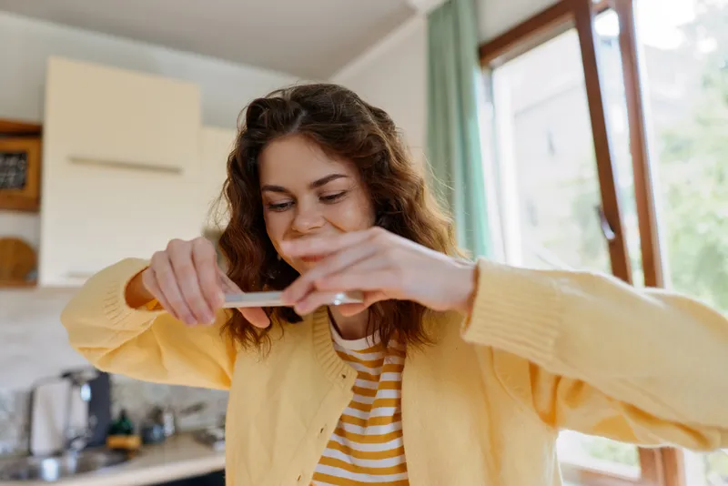 A young woman with curly hair in a yellow cardigan sits in a bright kitchen at home, gently tapping her smartphone screen and smiling warmly at a moment of everyday connection.
