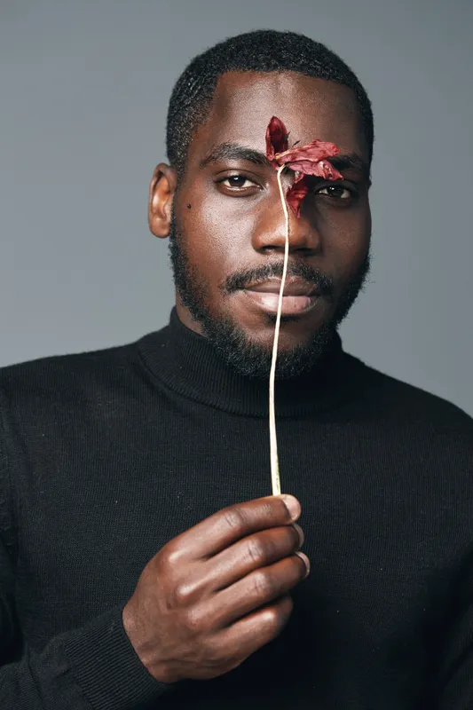 African man with dark skin holding dried flower in front of face in creative conceptual visual style. Portrait with black turtleneck and neutral background for artistic expression and emotion.