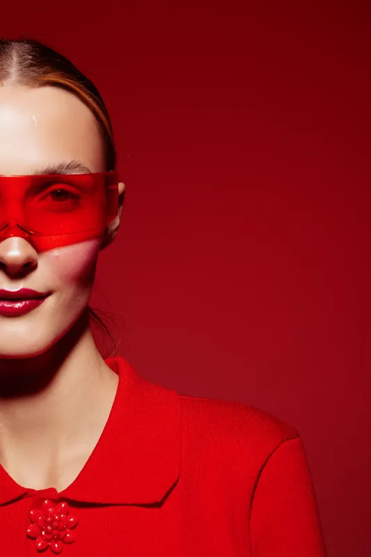 Young woman in red sweater and red translucent glasses poses against red background. Fashion portrait with bold color contrast and modern style concept.