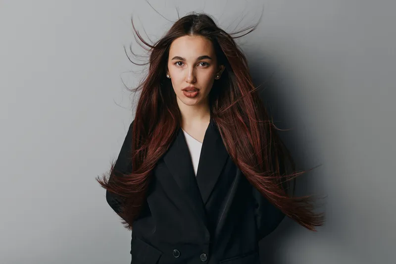 Woman with long brown hair wearing black blazer and white shirt posing against gray background. Portrait of confident female with natural makeup and serious expression in studio lighting.