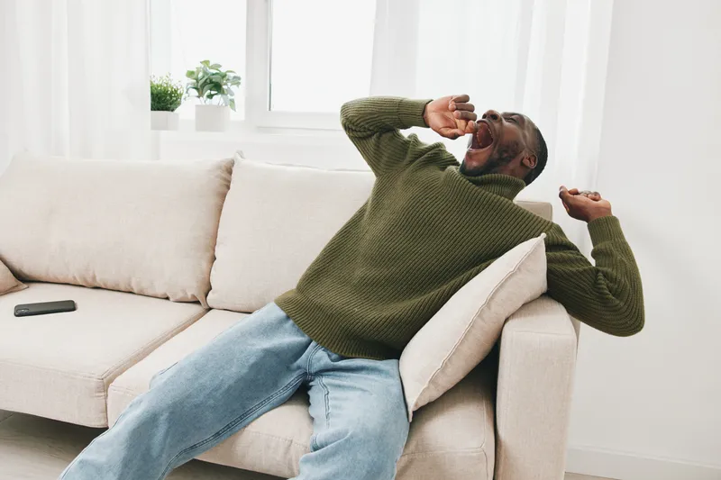 A relaxed Black man yawning while sitting on a beige couch, wearing a green sweater and light jeans, capturing a cozy and laid back atmosphere in a modern living room