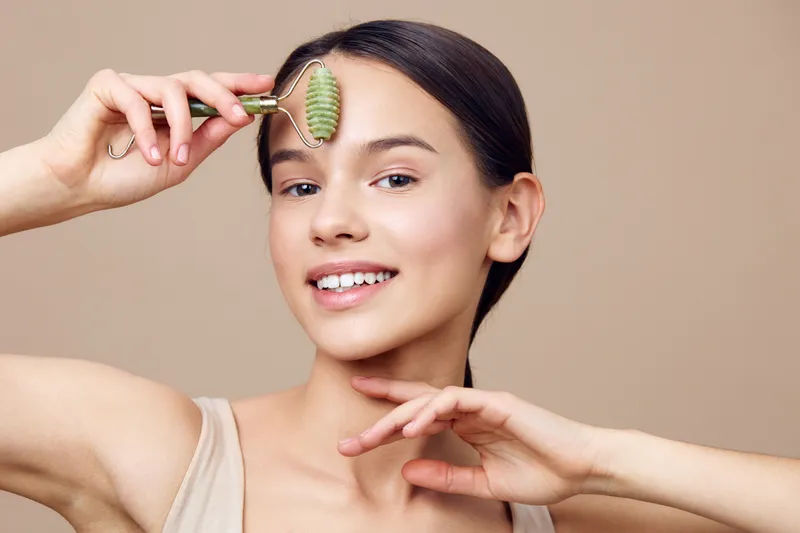 Young woman using a jade roller for skincare, showcasing a natural glow, light background, emphasizing beauty and wellness