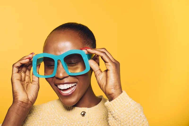 Smiling black African woman with dark skin wearing oversized blue glasses and yellow sweater stands against bright yellow background. Concept for fashion, joy, and vibrant style.