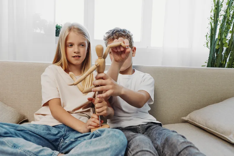 Two children sitting on a sofa play with a wooden mannequin figure in a bright living room with natural light and indoor plants. Concept of childhood, creativity, and family time.