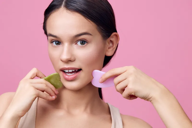 Young woman demonstrating facial skincare with jade and rose quartz tools, emphasizing beauty and wellness against a vibrant pink background