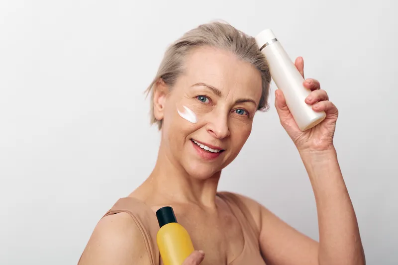 Smiling middle aged woman with short blonde hair applies moisturizing cream on cheek holding cosmetic bottles. Concept of skincare routine, anti aging care, wellness, and natural beauty at home.