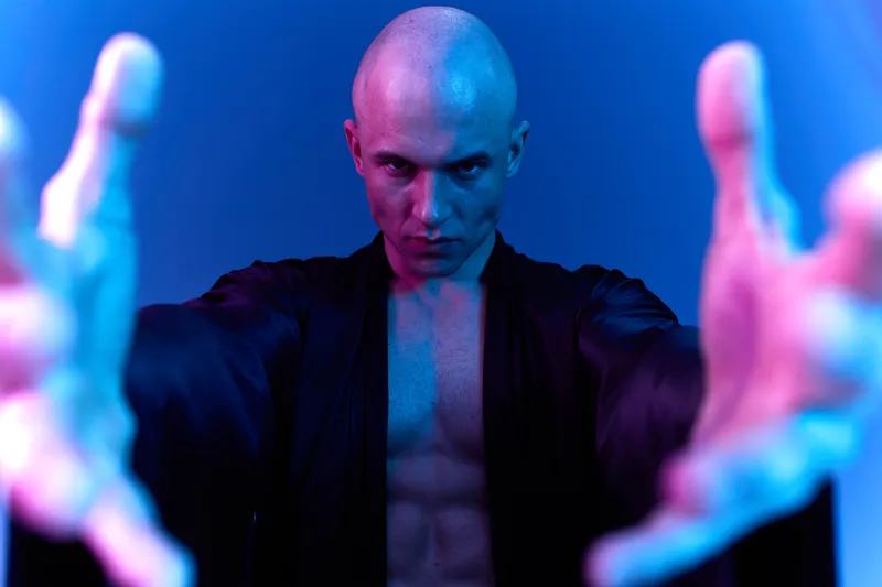Intense man with a bald head, wearing a dark robe, showcasing a dramatic expression and extended arms. The image captures a unique, moody atmosphere filled with blue and purple light.