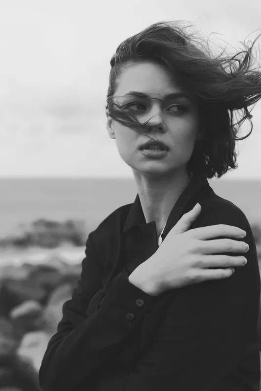 Windy coastal scene with a woman in dark clothing, hair swept by the breeze, standing along a rocky shore in a moody monochrome, capturing dramatic, editorial fashion and natural emotion by the sea