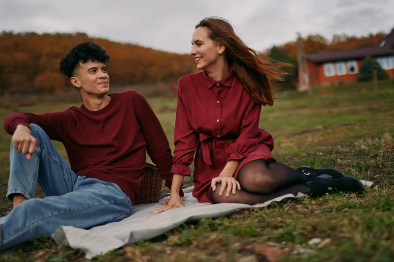 A young mixed couple sitting on a blanket in a rural hillside, sharing a candid smile and genuine conversation that conveys authenticity and credibility in a relaxed outdoor lifestyle portrait.