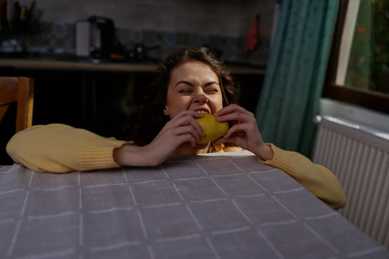 Young woman with brunette hair in a yellow sweater leans over a kitchen table, biting into a yellow fruit with a relaxed smile, capturing a cozy home lifestyle moment.