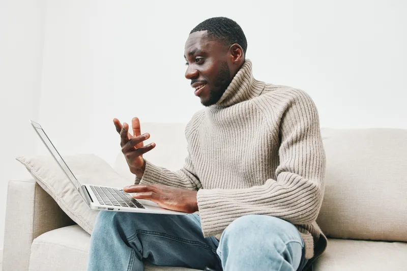 Happy African man in cozy sweater using laptop for online communication at home, enjoying comfortable lifestyle and technology connection
