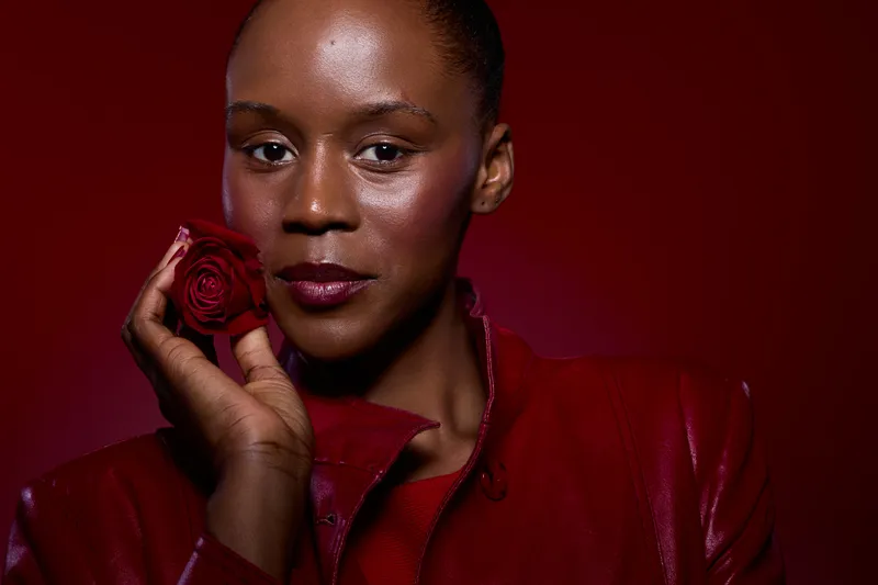 Close-up portrait of a confident dark skinned African woman holding a red rose near her face. She wears a red leather jacket with a matching red background. Concept of beauty and elegance.