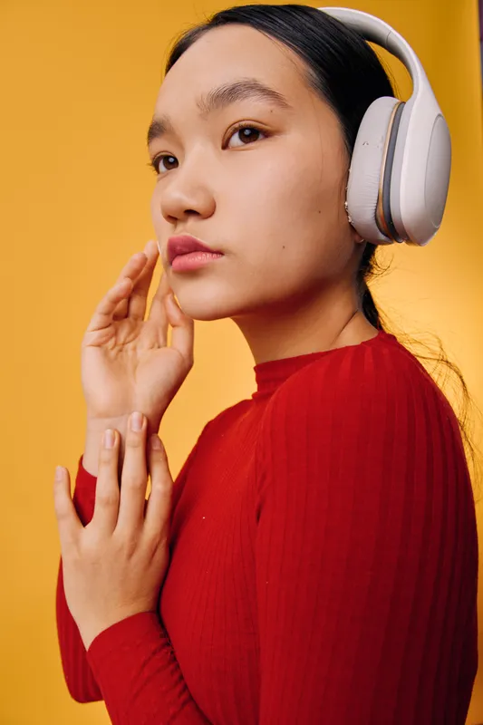 Young woman wearing wireless headphones listening to music or podcast with thoughtful expression on yellow background. Closeup portrait of female in red sweater enjoying sound experience.