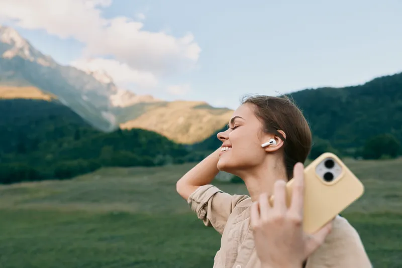 Woman outdoors lifestyle portrait, smiling as she takes a selfie with a phone and earbuds, green field and distant mountains in the background, peaceful moment outdoors