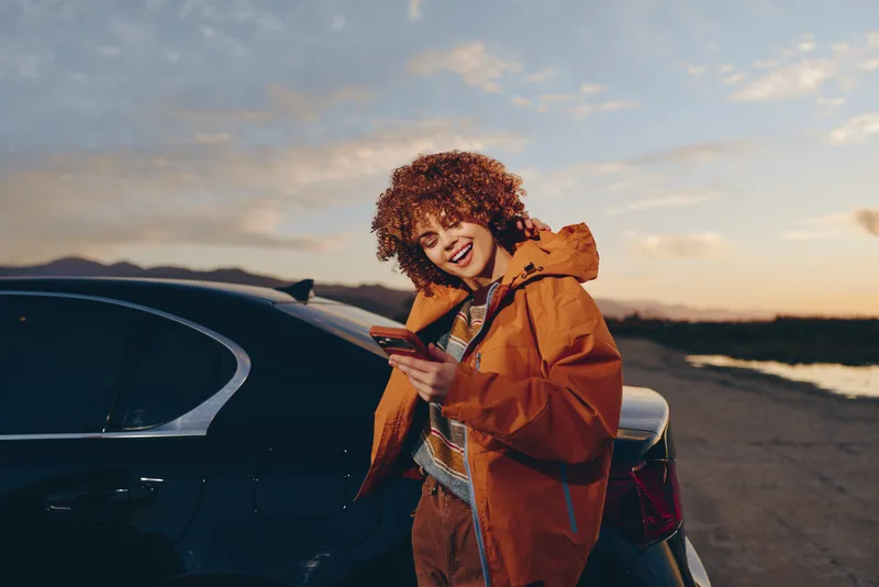 Woman in orange jacket near a black car on a seaside road at sunset, holding a smartphone and smiling, lifestyle travel scene, warm evening light, outdoors