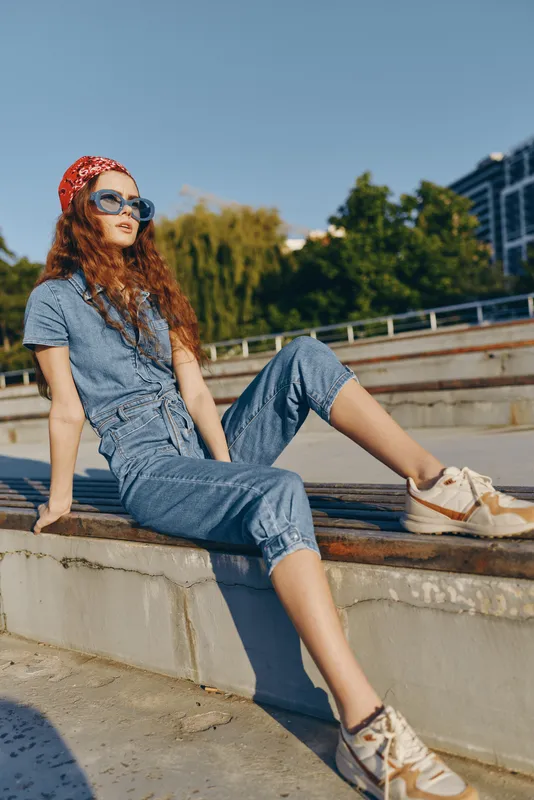 Lifestyle woman in modern boho-western denim street style sitting outdoors, wearing sunglasses and sneakers, film color tones, relaxed urban fashion vibe.
