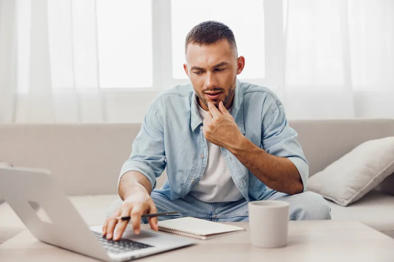 Thoughtful man working on a laptop at home, wearing casual denim attire, focused on tasks. Soft natural light enhances the home office ambiance, promoting productivity and creativity.