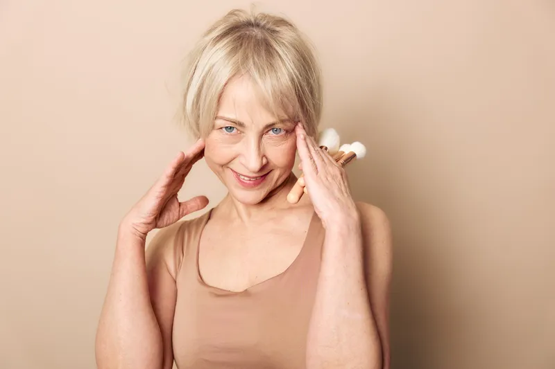 Smiling mature woman with blonde hair touches face gently, wearing beige top against neutral background. Concept for beauty, aging, skin care, confidence, and self-love.