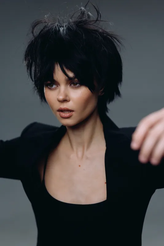 Young woman with messy black hair and stylish black outfit posing in a studio setting with a neutral gray background.