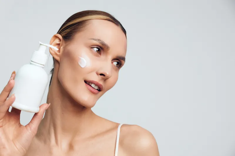 Young woman applies moisturizer from a pump bottle, close up portrait showing clear skin and soft makeup, skincare routine moment, minimal background, beauty care