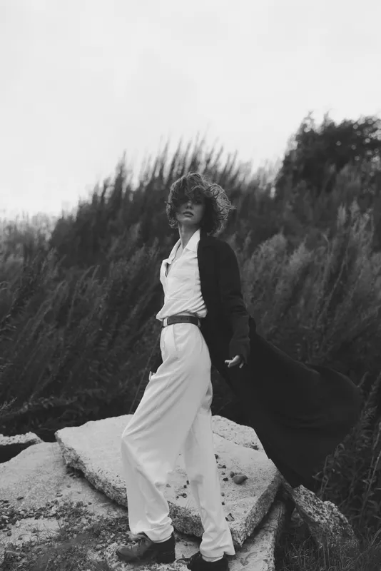 Fashion model in a flowing dress and long coat strikes a bold pose outdoors, wind in her hair, rocky terrain and tall grasses creating contrast in monochrome with mood.
