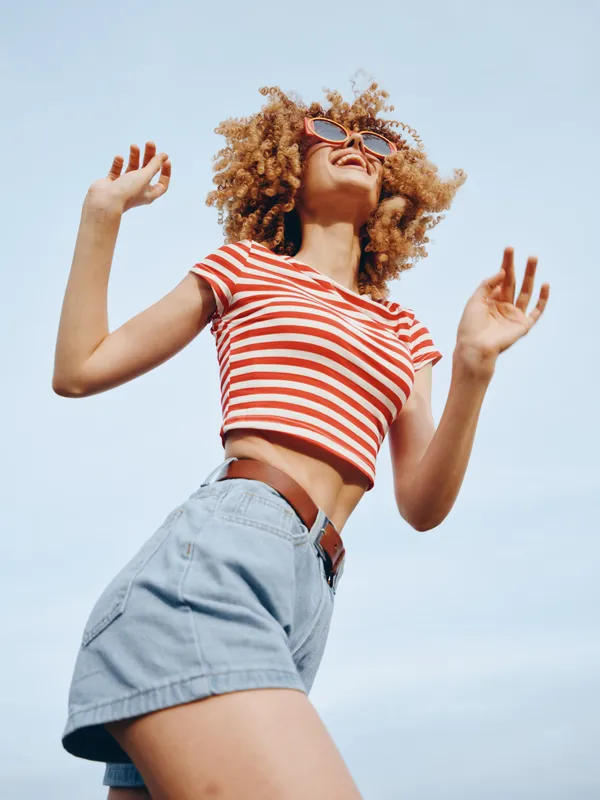 A joyful young woman with curly hair dances outdoors under a bright blue sky, wearing a red striped crop top and light denim shorts, captured from a low angle.