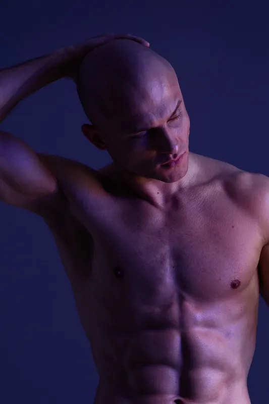 Powerful man posing in a dramatic light, showcasing his toned physique and strong features. The image captures an intense and artistic mood, celebrating strength.