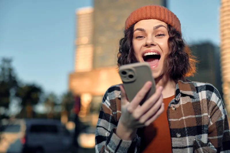Woman smartphone laughter in urban street, candid beanie portrait with authenticity and lifestyle mood, golden hour glow, mindful living and emotional storytelling in warm sunset light.