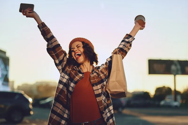 woman joy coffee shopping portrait urban golden candid authenticity mindful living emotional storytelling Young woman raises coffee cup and shopping bag in golden hour glow, smiling in a candid