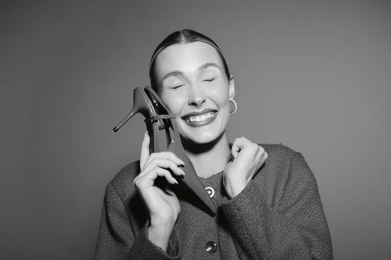 Smiling Caucasian woman with pale skin holding black high heel shoe close to face, eyes closed, wearing dark sweater and hoop earrings. Concept of fashion, happiness, beauty and style.