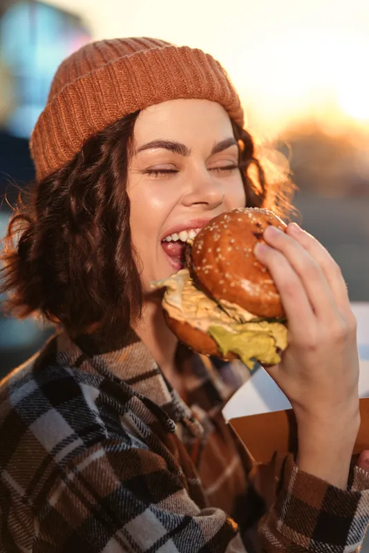woman burger eating smile beanie plaid golden hour candid outdoor happy portrait of a young urban foodie biting a sesame bun at sunset, authenticity, candid lifestyle, golden hour glow, mindful