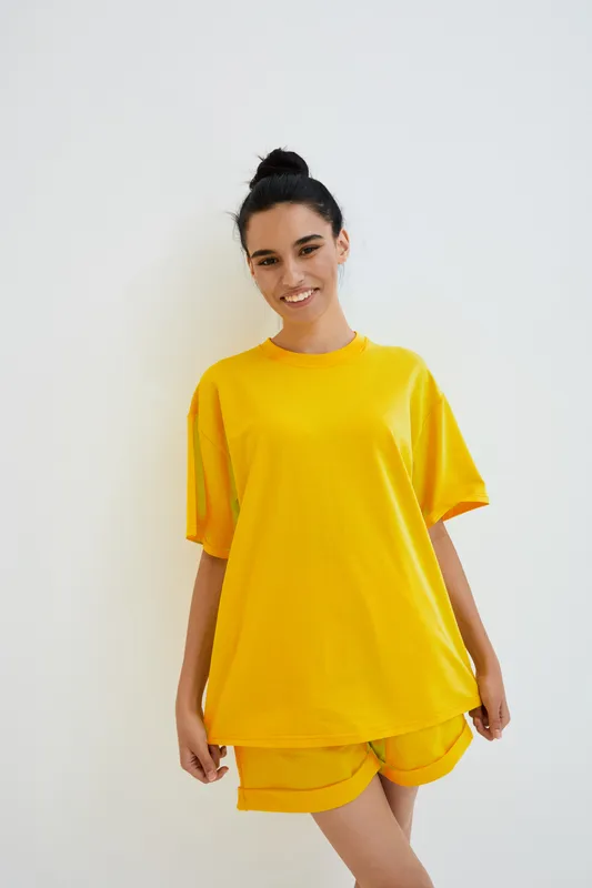 young woman in bright yellow casual outfit smiling confidently with natural makeup and hair tied up, standing against plain white background with relaxed mood