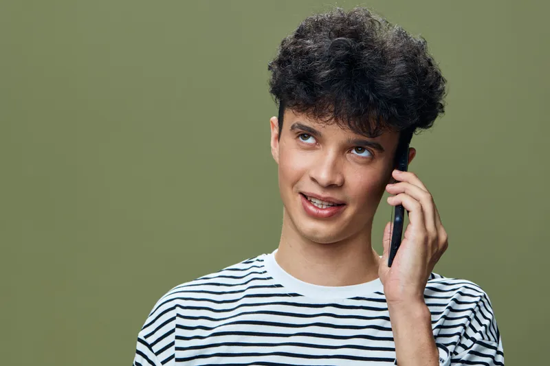 young man with curly hair talking on smartphone, wearing striped shirt against olive background, casual tech communication portrait with attentive expression