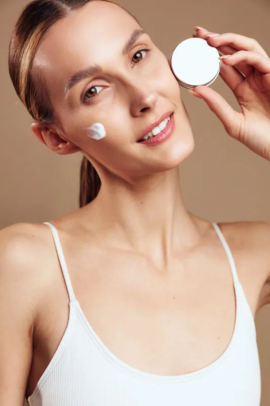 Young woman with natural skin applies face cream on cheek holds jar of moisturizer looks at camera with gentle smile isolated on beige background in white tank top