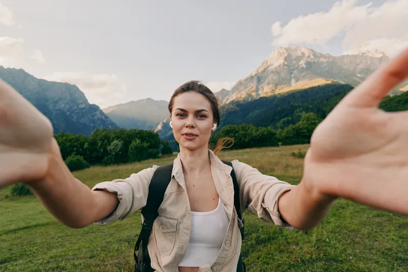 A young woman in casual clothes takes a selfie outdoors in a green meadow with mountains in the background, enjoying nature and an active lifestyle.