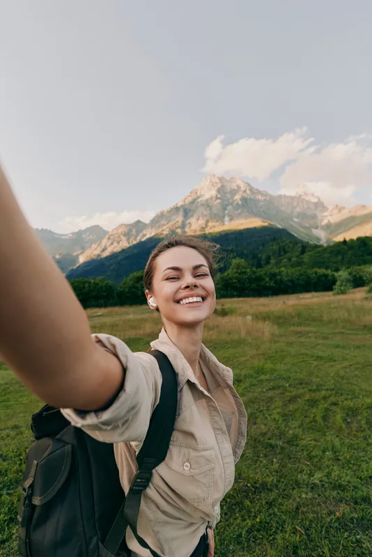 Smiling woman outdoors taking a selfie in a grassy meadow with distant mountains, capturing a lifestyle of travel, wellness, and nature, bright daylight and warm mood