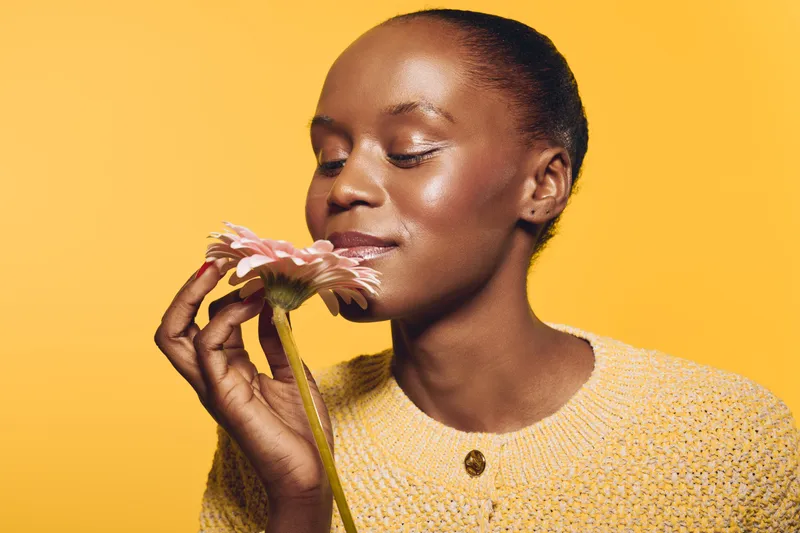 Close-up of an African woman with dark skin holding a pink flower near her face against a yellow background. Concept of natural beauty, floral scent, and peaceful moment.